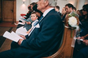 Während einer standesamtlichen Trauung versammeln sich die Gäste in den Kirchenbänken; ein Junge lächelt in die Kamera.