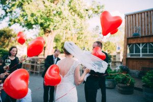 Brautpaar lächelt Hand in Hand mit Gästen und roten Luftballons bei Hochzeit Gut Knittkuhle Düsseldorf.