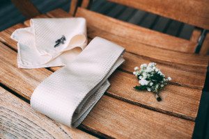 Cremefarbene Krawatte, Einstecktuch, Manschettenknöpfe und Blumenboutonniere auf einem Stuhl – Hochzeitsreportage NRW.