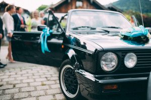 Ein schwarzes Hochzeitsauto mit blauen Bändern auf einer Kölner Straße, die Gäste unterhalten sich nach der standesamtlichen Trauung.