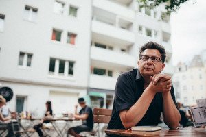 Hochzeitsgast mit Brille sitzt entspannt an einem gemütlichen Cafétisch in Köln und lächelt sanft.