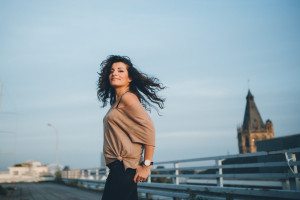 Braut mit lockigem Haar lächelt über ihre Schulter auf einer sonnigen Promenade in Köln, hinter ihr der Kölner Dom.