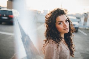 Braut mit wallenden Locken steht auf einem sonnenbeschienenen Kölner Parkplatz und lächelt sanft in die Kamera. Hochzeitsfotografin Köln.