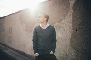 Ein Mann im blauen Pullover lehnt entspannt an einer sonnenbeschienenen Wand in Köln, Portrait von Hochzeitsfotografin.