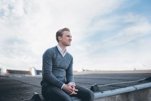 Ein Mann im grauen Pullover sitzt entspannt auf einem Dach – moderne Portraitfotos in Köln.