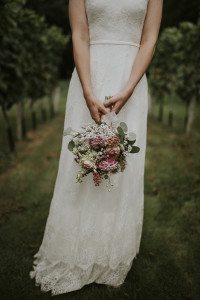 Eine Braut im Spitzenkleid lächelt sanft mit ihrem Blumenstrauß auf der Hochzeit WinselerHof – Hochzeitsfotografin Köln.