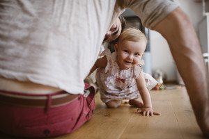 Ein Baby in einem leichten Kleid krabbelt und lächelt während Familienfotos Köln auf einem Holzboden.