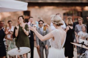 Eine Braut auf Gut Hohenholz hebt lächelnd zwei Bier, während sich Freunde zu einer entspannten Hochzeitsreportage NRW treffen.