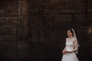 Eine Braut im weißen Kleid lächelt entspannt vor dunkler Holzwand – Hochzeitsreportage NRW.
