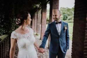 Brautpaar Hand in Hand unter einem Backsteinbogen, umgeben von Grün – Hochzeitsfotografin Köln.