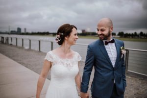 Das Brautpaar geht Hand in Hand am Rhein entlang, festgehalten von Hochzeitsfotografin Köln.