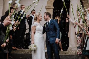 Braut und Bräutigam lächeln sich draußen an, umgeben von Gästen mit weißen Blumen – authentisch festgehalten von einer Hochzeitsfotografin Köln.