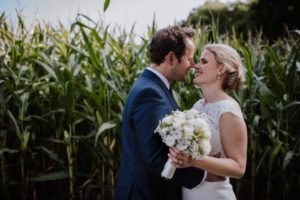 Brautpaar lächelt innig vor einem Kornfeld mit weißem Brautstrauß, Hochzeitsfotografin Köln.