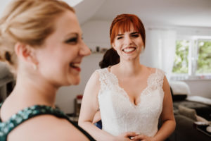 Braut im Spitzenkleid lächelt herzlich; Freundin im grünen Kleid steht daneben während der Hochzeitsreportage NRW.