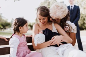 Eine Braut hält ihr Baby auf der Bank, umgeben von Familie – natürliche Hochzeitsfotografie Bonn.