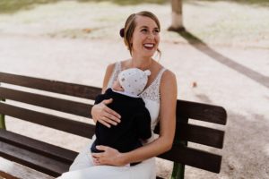Braut in weiß auf einer Bank, lächelt liebevoll mit Baby im Arm – Hochzeitsreportage NRW.