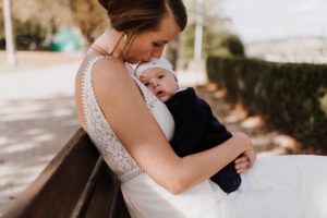 Eine Braut im Spitzenkleid hält ihr Baby auf der Bank – liebevoller Moment einer Hochzeitsreportage NRW.