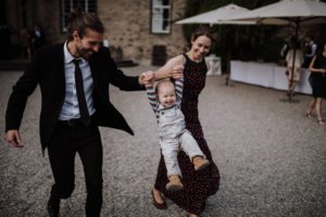 Eltern wiegen ihr lächelndes Kind an den Händen vor einem Steinveranstaltungsort in NRW, Lachen erfüllt den Tag.