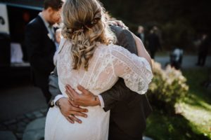 Braut im Spitzenkleid umarmt eine Vertraute im Sonnenlicht – Hochzeitsfotografin Köln hält den Moment fest.
