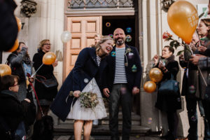 Neuer Alternativtext: Brautpaar lacht mit Freunden vor dem Standesamt in NRW, umgeben von Luftballons und Seifenblasen.