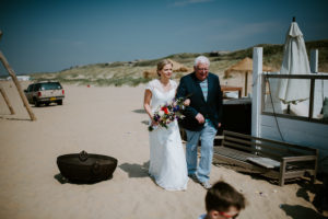 Braut im weißen Kleid mit Brautstrauß geht am Strand neben ihrem Vater – Hochzeitsfotografin Köln.