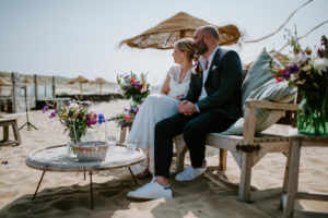Brautpaar sitzt entspannt am Strand auf einer Holzbank, umgeben von Blumen – Hochzeitsfotografin Köln.