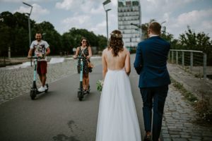 Braut und Bräutigam schlendern im Sonnenschein, während zwei E-Scooter an ihnen vorbeifahren. Hochzeitsreportage NRW.