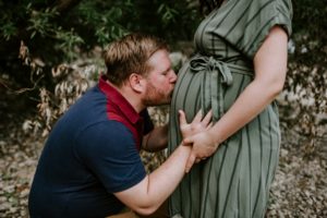 Ein Paar im Grünen, er küsst liebevoll ihren Babybauch – festgehalten von eurer Hochzeitsfotografin Köln.