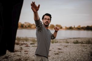 Bräutigam im gestreiften Hemd streckt sich am Flussufer entgegen – authentische Hochzeitsmomente in NRW.