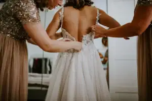 Zwei Freundinnen helfen einer Braut beim Schließen ihres Spitzenkleids – Hochzeitsfotografin Köln.